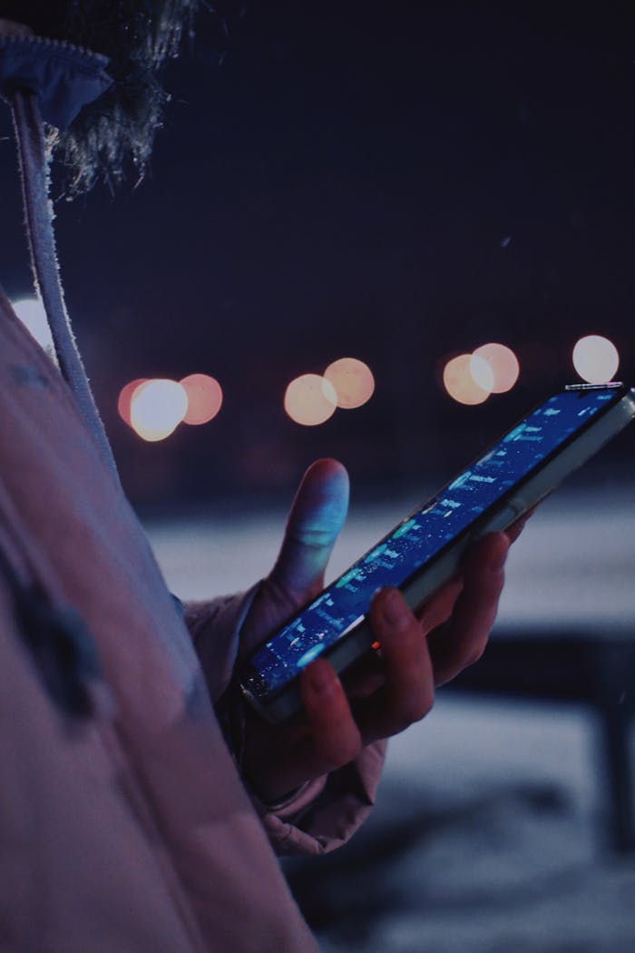 Close-up of hand holding smartphone illuminated by city lights during nighttime.